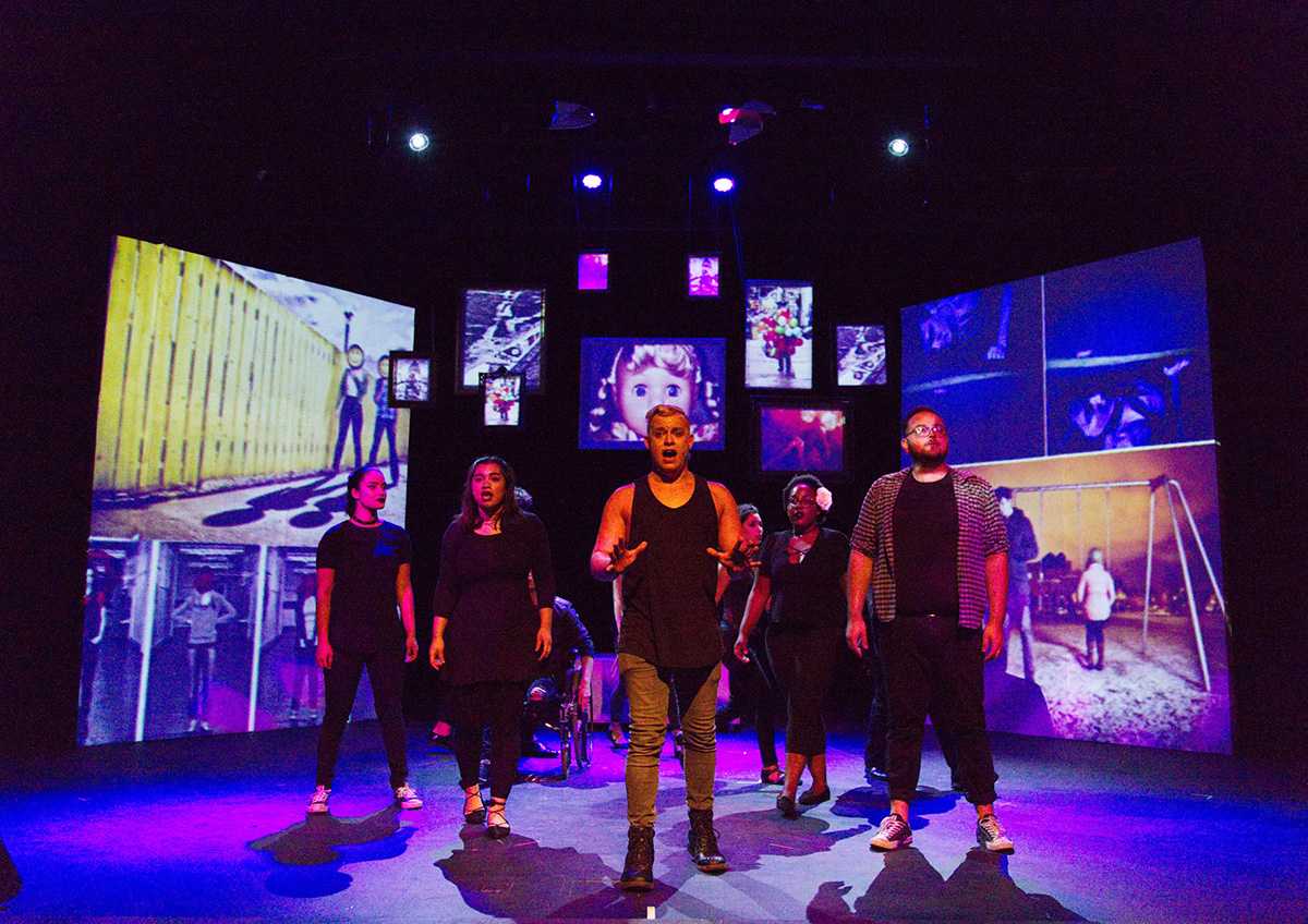 Photograph of cast on stage with projection screens behind them.