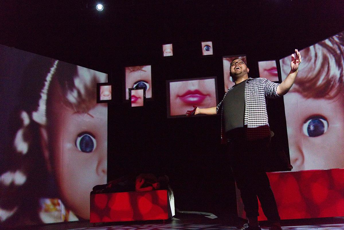 Man singing on stage in 35MM: A Musical Exhibition with images of doll face behind him.