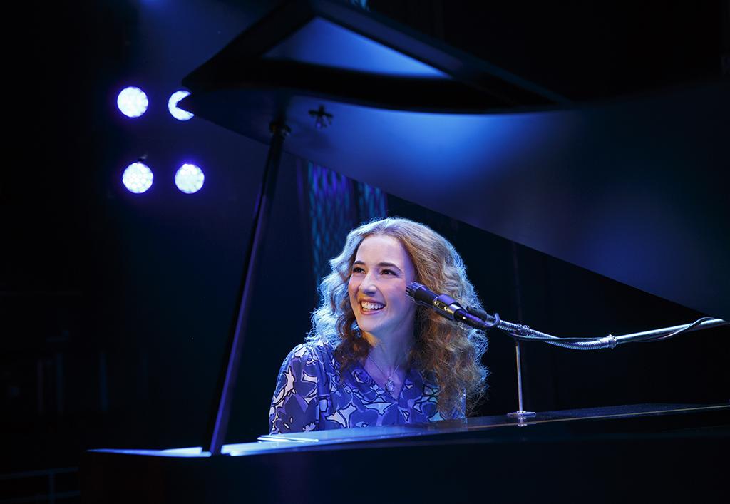 Actress playing Carole King sitting at piano.